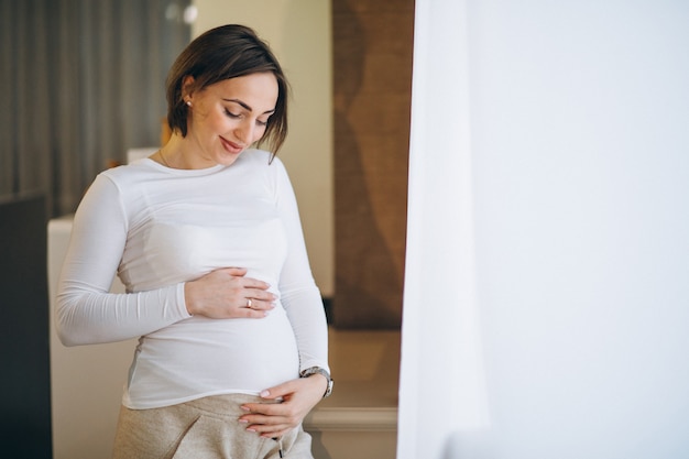 自宅の窓のそばに立っている若い妊娠中の女性