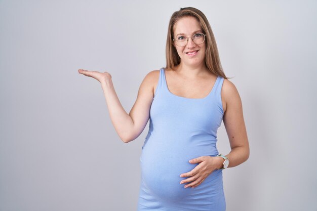 Молодая беременная женщина, стоящая на белом фоне, весело улыбается и показывает ладонью, глядя в камеру.