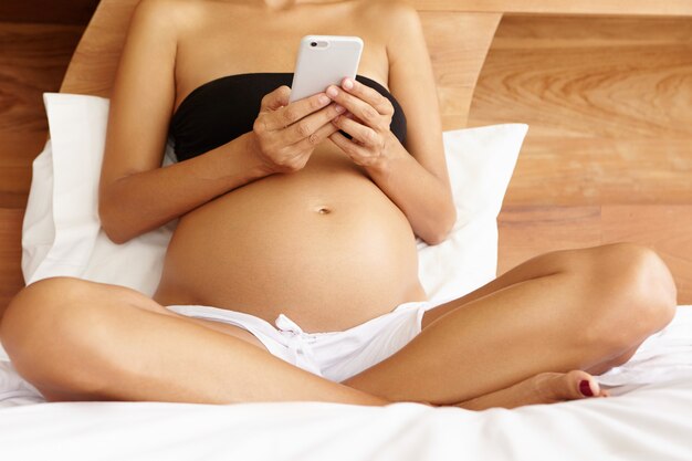 Молодая беременная женщина в нижнем белье расслабляется на кровати утром, просматривая фотографии в Интернете с помощью электронного устройства. Обрезанный вид женщины, ожидающей ребенка, читающей электронную книгу на смартфоне