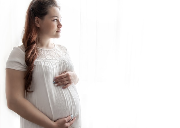 A young pregnant woman in a white T-shirt looks thoughtfully out the window.