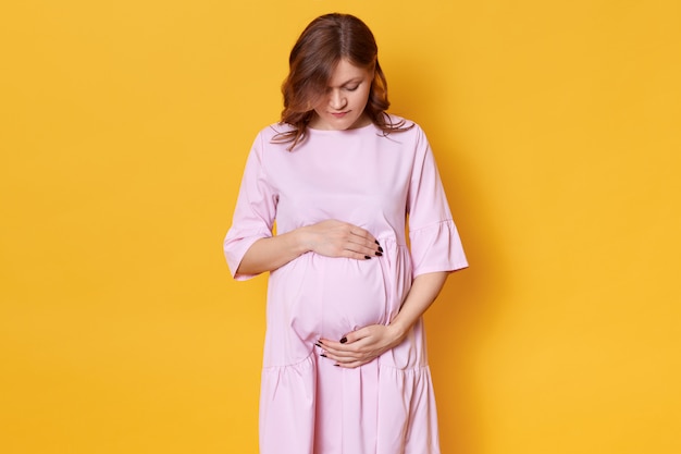 Young pregnant woman with brown hair, in elegant rose dress standing with hands on her belly in front of yellow, looking down on her abdomen, has dark manicure.