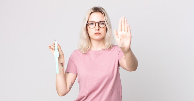 Young pretty albino woman looking serious showing open palm making stop gesture holding a watch clock