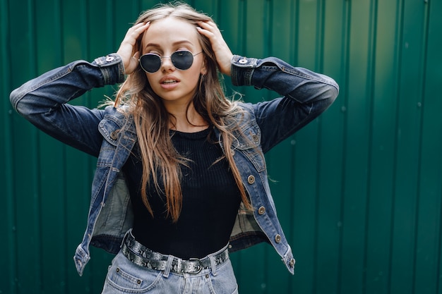 Free photo young pretty attractive girl in denim clothes in glasses on a simple dark green wall with place for text.