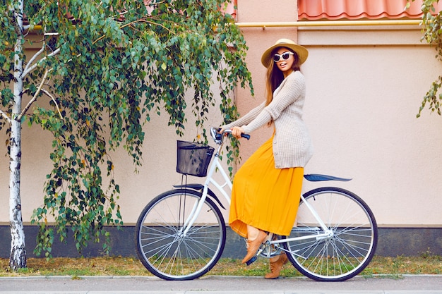 Молодая довольно красивая женщина, катающаяся на своем белом ретро-хипстерском велосипеде, в стильных винтажных солнцезащитных очках и соломенной шляпе, модный осенний портрет элегантной дамы с удовольствием на открытом воздухе.