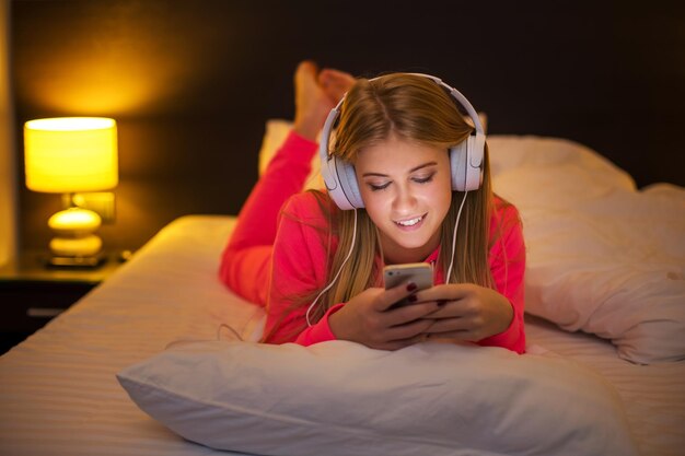 自宅のベッドに横たわっているスマートフォンからヘッドセットで音楽を聴いて笑っている若いきれいなブロンドの女性