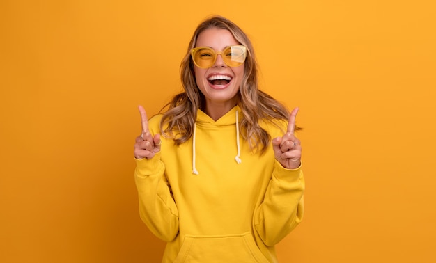 Free photo young pretty blonde woman cute face expression posing in yellow hoodie on yellow bright isolated