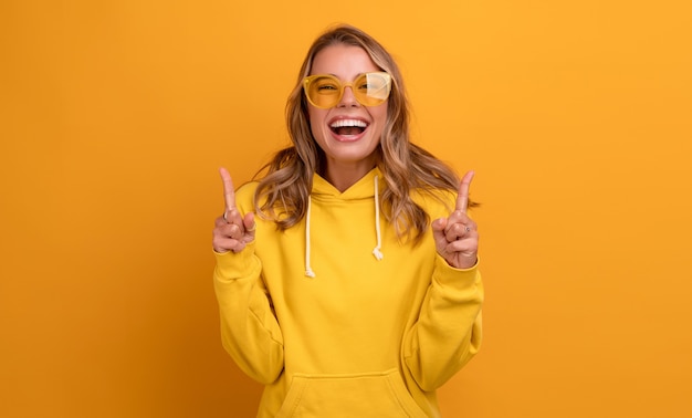 Young pretty blonde woman cute face expression posing in yellow hoodie on yellow bright isolated