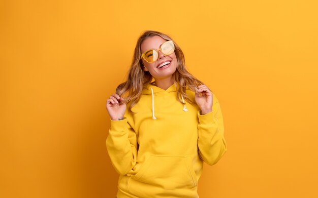 Young pretty blonde woman cute face expression posing in yellow hoodie on yellow bright isolated