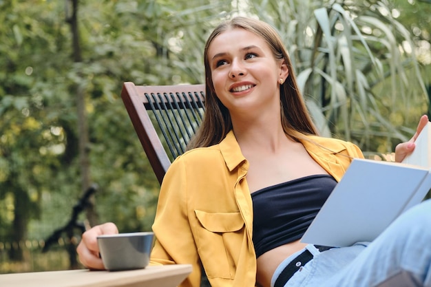 Young pretty brown haired teenage girl in yellow shirt and top happily sitting with book and cup of coffee on wooden deck chair in city park