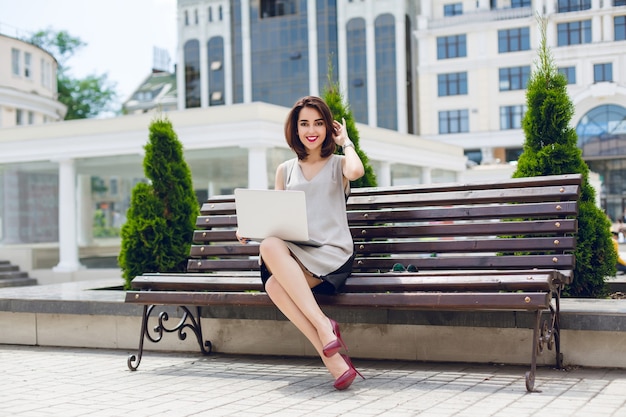 Молодая красивая деловая женщина брюнетка сидит на скамейке в городе. На ней серо-черное платье, туфли на высоких каблуках и бордовые губы.