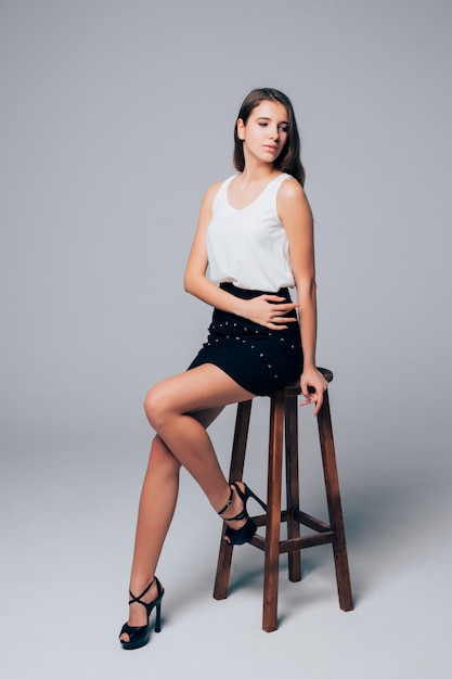 Young pretty brunette in short black skirt sit on high wood chair isolated on white