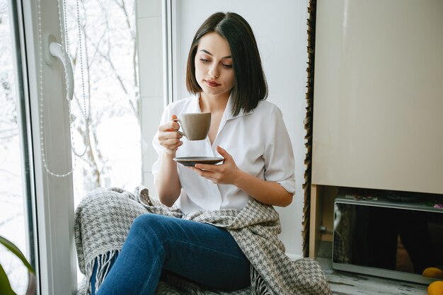 コーヒーを持つ台所の窓に座っている若くて美しいブルネット