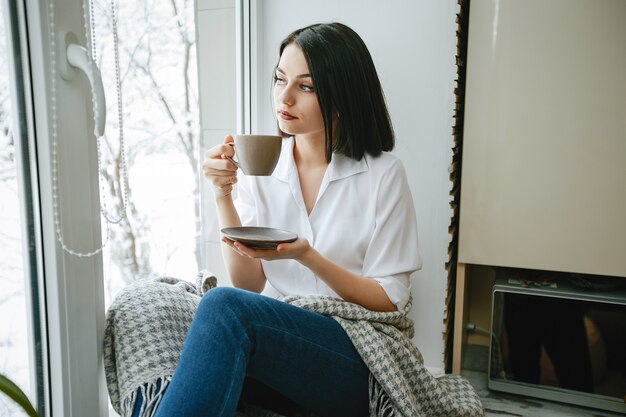 молодая и красивая брюнетка, сидя у окна на кухне с кофе