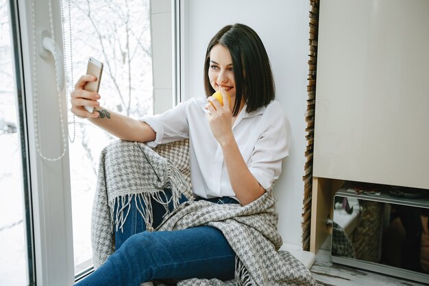 young and pretty brunette sitting by the window in the kitchen with lemon and phone