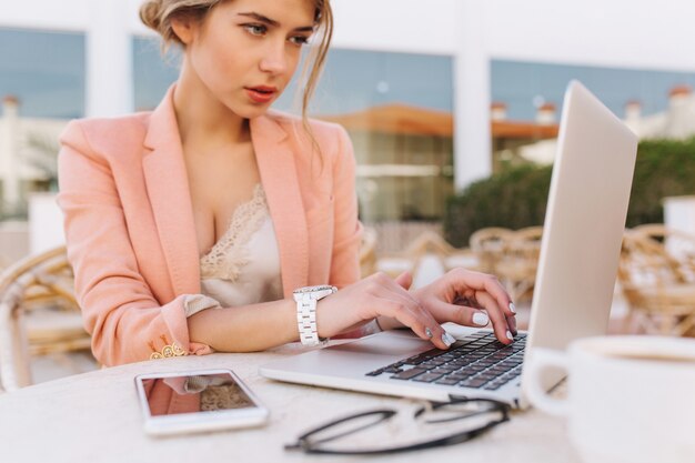 Молодая красивая деловая женщина, работающая на ноутбуке в уличном кафе, набрав на клавиатуре, умная дама серьезно смотрит на экран. Смартфон и очки на столе. Одет в стильный розовый пиджак, белые часы.