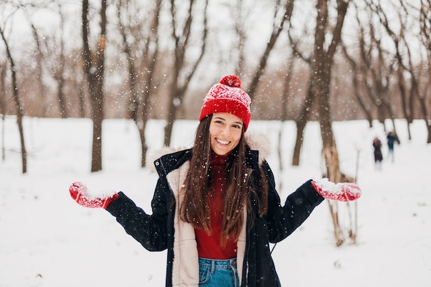 Giovane donna felice sorridente abbastanza candida in guanti rossi e cappello che indossa cappotto nero che cammina giocando nel parco nella neve in vestiti caldi, divertendosi