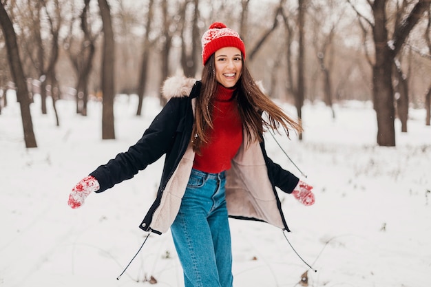 Giovane donna felice sorridente abbastanza candida in guanti rossi e cappello che indossa cappotto nero che cammina giocando nel parco nella neve in vestiti caldi, divertendosi