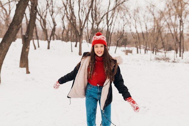 赤いミトンと帽子をかぶった若いかなり率直な笑顔の幸せな女性は、暖かい服を着て雪の中で公園で遊んで歩いて、楽しんで