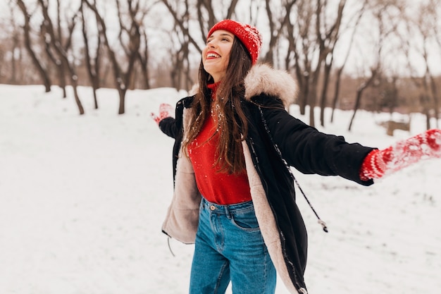 Young pretty candid smiling happy woman in red mittens and hat wearing black coat walking playing in park in snow in warm clothes, having fun
