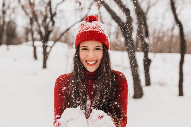 Young pretty candid smiling happy woman in red mittens and hat wearing knitted sweater walking playing in park in snow, warm clothes, having fun