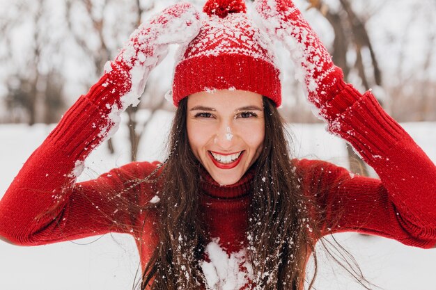 Young pretty candid smiling happy woman in red mittens and hat wearing knitted sweater walking playing in park in snow, warm clothes, having fun