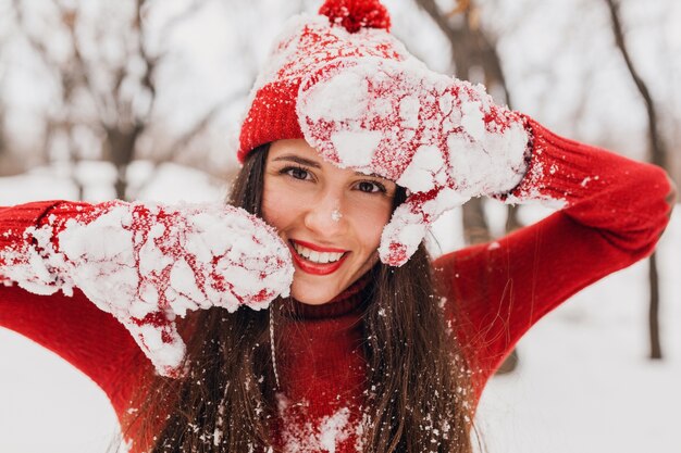 Young pretty candid smiling happy woman in red mittens and hat wearing knitted sweater walking playing in park in snow, warm clothes, having fun
