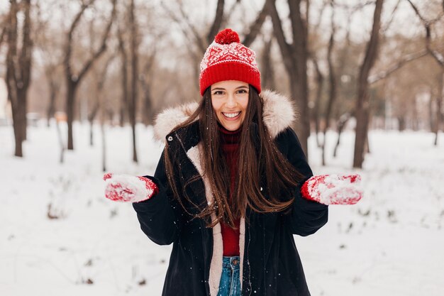 Young pretty candid smiling happy woman in red mittens and knitted hat wearing black coat walking playing in park in snow, warm clothes, having fun