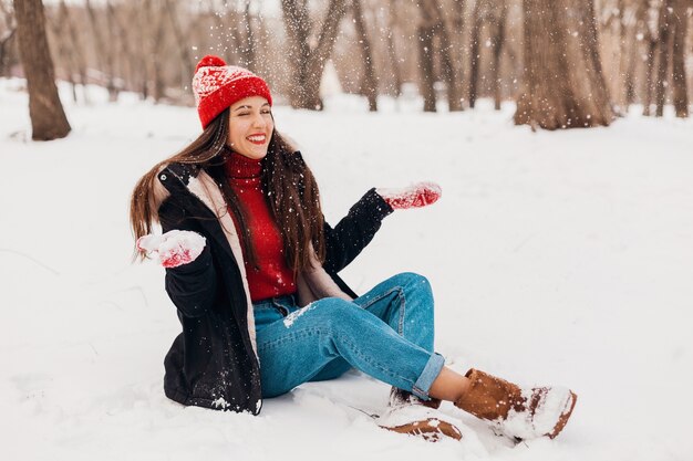 Молодая довольно откровенная улыбающаяся счастливая женщина в красных рукавицах и вязаной шапке в черном пальто гуляет, играя в парке в снегу, теплой одежде, веселится