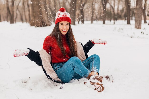 Young pretty candid smiling happy woman in red mittens and knitted hat wearing black coat walking playing in park in snow, warm clothes, having fun