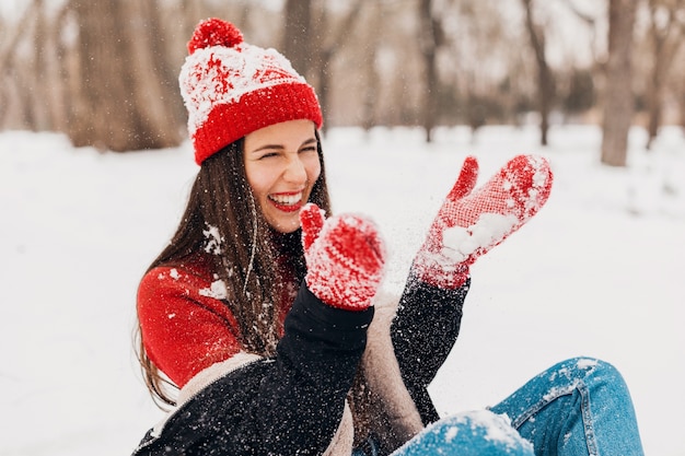 Young pretty candid smiling happy woman in red mittens and knitted hat wearing black coat walking playing in park in snow, warm clothes, having fun