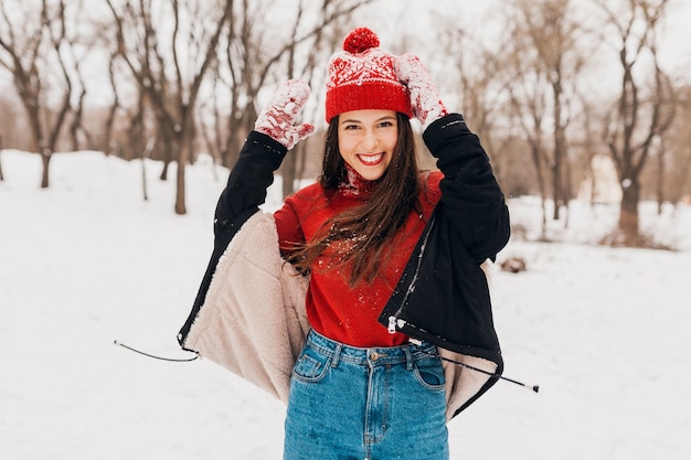 Young pretty candid smiling happy woman in red mittens and knitted hat wearing black coat walking playing in park in snow, warm clothes, having fun