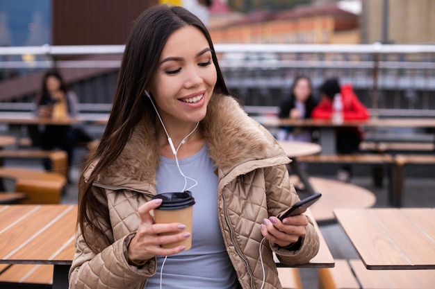 屋外の携帯電話でイヤホンで楽しく音楽を聴きに行くためにコーヒーとダウンジャケットの若いかなりカジュアルな女性