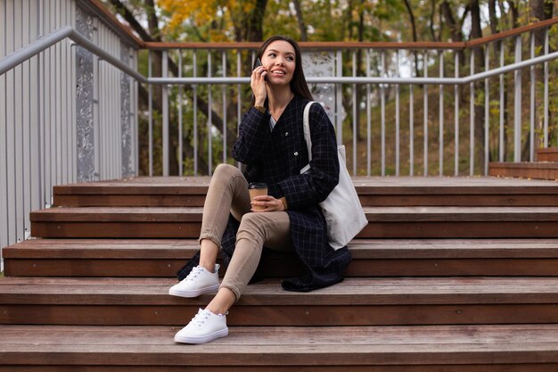 Young pretty casual woman with coffee to go happily talking on cellphone in city park