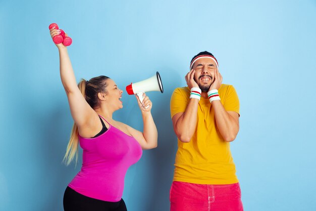 Young pretty caucasian couple in bright clothes training on blue background Concept of sport, human emotions, expression, healthy lifestyle, relation, family. He's scared, she's calling in mouthpeace.