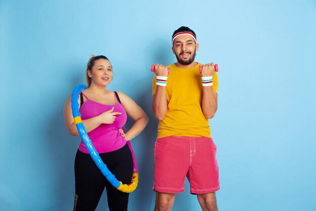 Young pretty caucasian couple in bright clothes training on blue background Concept of sport, human emotions, expression, healthy lifestyle, relation, family. Practicing with hoop and weights.