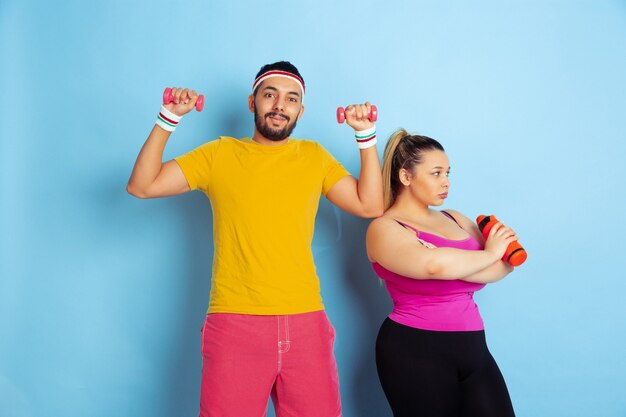 Young pretty caucasian couple in bright clothes training on blue background Concept of sport, human emotions, expression, healthy lifestyle, relation, family. Training with weights, have fun.
