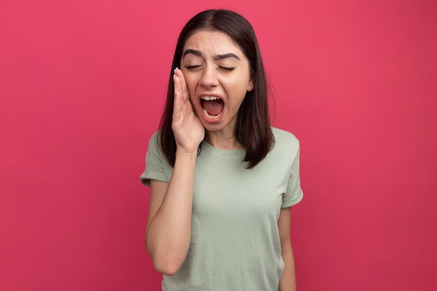 young pretty caucasian girl keeping hand near mouth calling out loud to someone with closed eyes isolated on pink wall with copy space