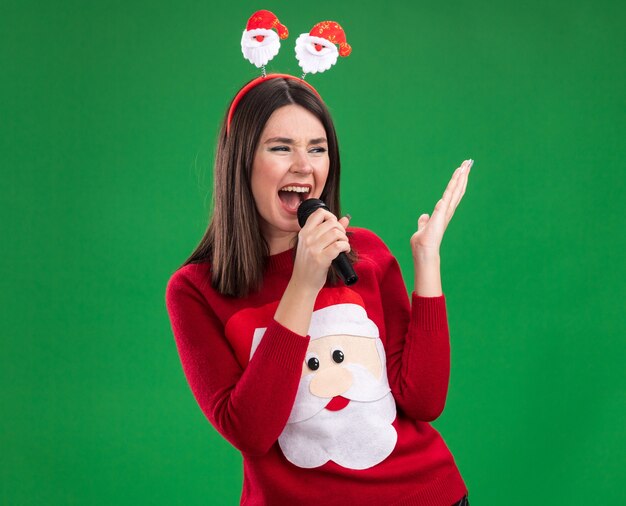 Young pretty caucasian girl wearing santa claus sweater and headband holding microphone looking at side keeping hand in air singing isolated on green wall with copy space