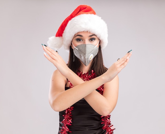 Young pretty caucasian girl wearing santa hat and protective mask tinsel garland around neck looking at camera doing no gesture isolated on white background