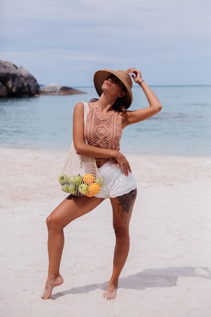 Young pretty caucasian tanned fit woman in knitted clothes on tropical beach wearing straw hat holding ecology reusable string bag full of fruits apple orange grapes
