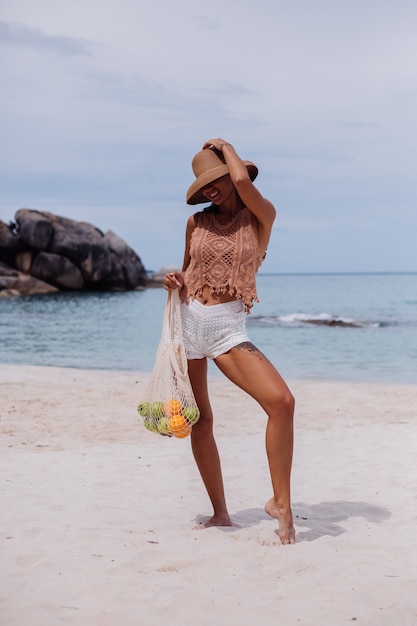 熱帯のビーチでニットの服を着た若いかなり白人の日焼けしたフィットの女性は、果物のリンゴオレンジブドウでいっぱいのエコロジー再利用可能なストリングバッグを保持している麦わら帽子を着ています