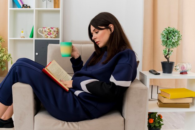 脚に本に触れて本を読んでカップを保持しているデザインのリビングルームの肘掛け椅子に座っている若いかなり白人女性
