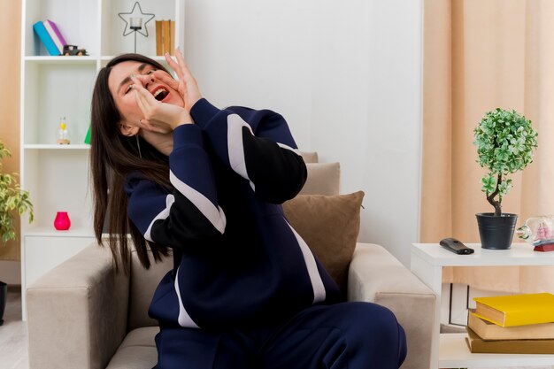 Young pretty caucasian woman sitting on armchair in designed living room keeping hands around mouth calling out loud to someone with closed eyes