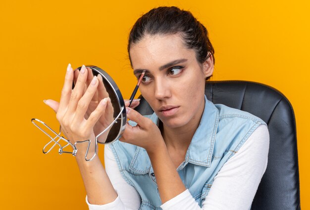 Young pretty caucasian woman sitting at table with makeup tools applying eyeshadow holding and looking at mirror isolated on orange wall with copy space