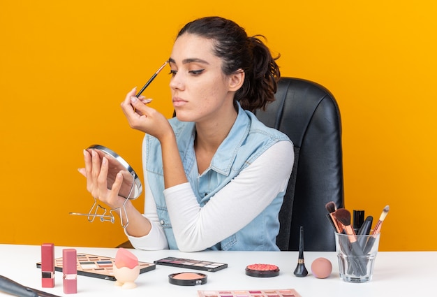 Молодая красивая кавказская женщина сидит за столом с инструментами для макияжа, применяя тени для век с кистью для макияжа, глядя в зеркало