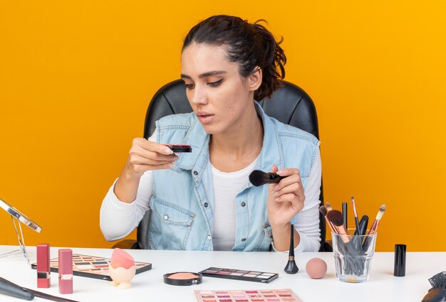 Молодая красивая кавказская женщина сидит за столом с инструментами для макияжа, держа кисть для макияжа и глядя на румянец