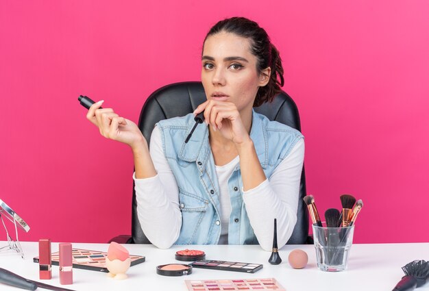 Young pretty caucasian woman sitting at table with makeup tools holding mascara isolated on pink wall with copy space