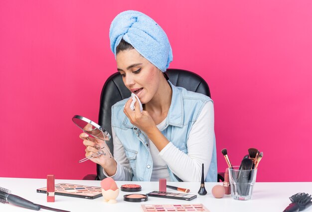 Young pretty caucasian woman with wrapped hair in towel sitting at table with makeup tools holding and looking at mirror wiping her mouth with napkin isolated on pink wall with copy space