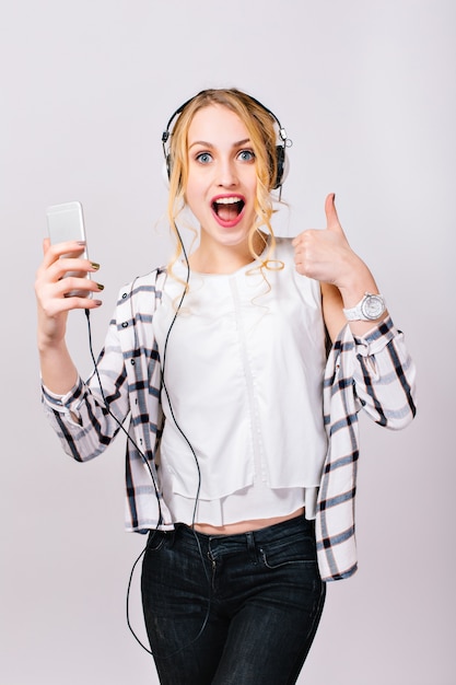 Young pretty cheerful girl with smartphone in hand. Merry gorgeous blonde in high spirits listening to music, showing Good sign and laughing. Exciting emotions. Satisfaction.