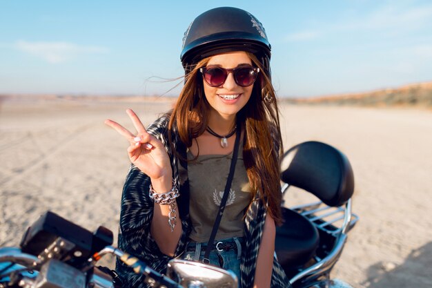 Young pretty cheerful  woman sitting on motorbike on the beach and show signs, wearing stylish crop top, shirts, have perfect fit slim tamed body and long hairs. Outdoor lifestyle portrait.